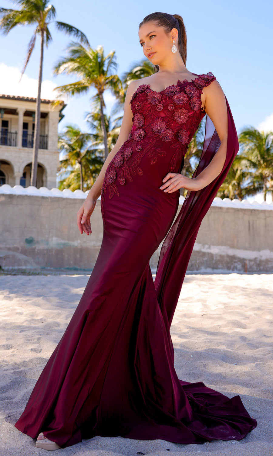 Woman posing on a sandy beach with palm trees and buildings behind her, wearing the One-Shoulder Long Formal Dress with Cape 388—a fitted, floor-length gown featuring a dramatic cape detail.
