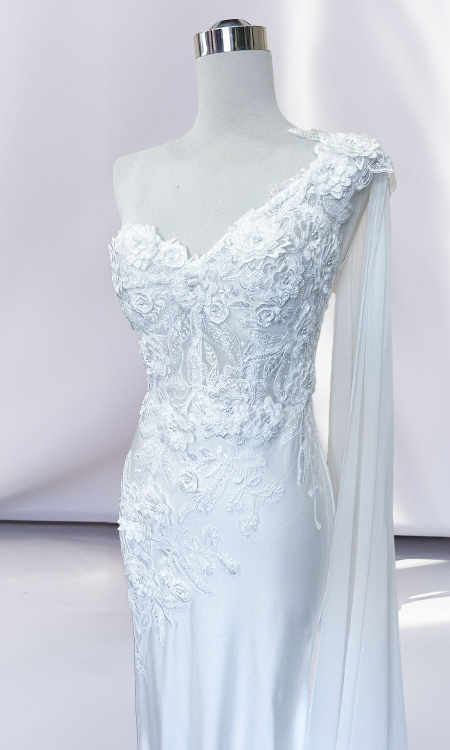 A white mannequin showcases the One-Shoulder Long Formal Dress with Cape 388, featuring a corset-style bodice, floral lace appliqué, and a draped sheer sleeve detail against a plain background.