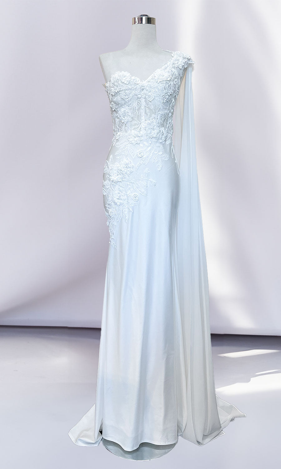 Displayed on a mannequin, the One-Shoulder Long Formal Dress with Cape 388 is a white, floor-length gown featuring a corset-style bodice, floral lace details, and a single draped shoulder sash against a plain background.