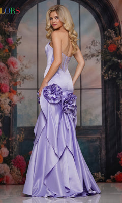 A woman wearing the Long Prom Dress: Colors Dress 3880, featuring 3D rosette appliques and bold floral skirt details, stands before a floral windowed backdrop, glancing over her shoulder.