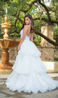 A woman wearing the Long Formal Dress 38813 by Ava Presley, featuring a white embroidered corset bodice, stands outdoors near a stone fountain with trees and greenery in the background, looking over her shoulder.