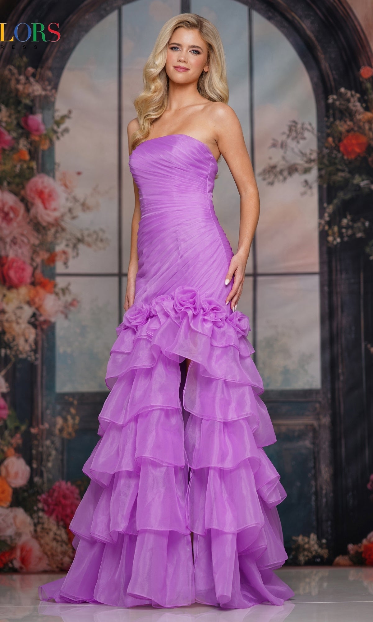 A woman in the Colors Dress 3882 long prom dress, featuring a strapless ruched bodice and ruffled lavender skirt, stands before a floral and arched window backdrop.