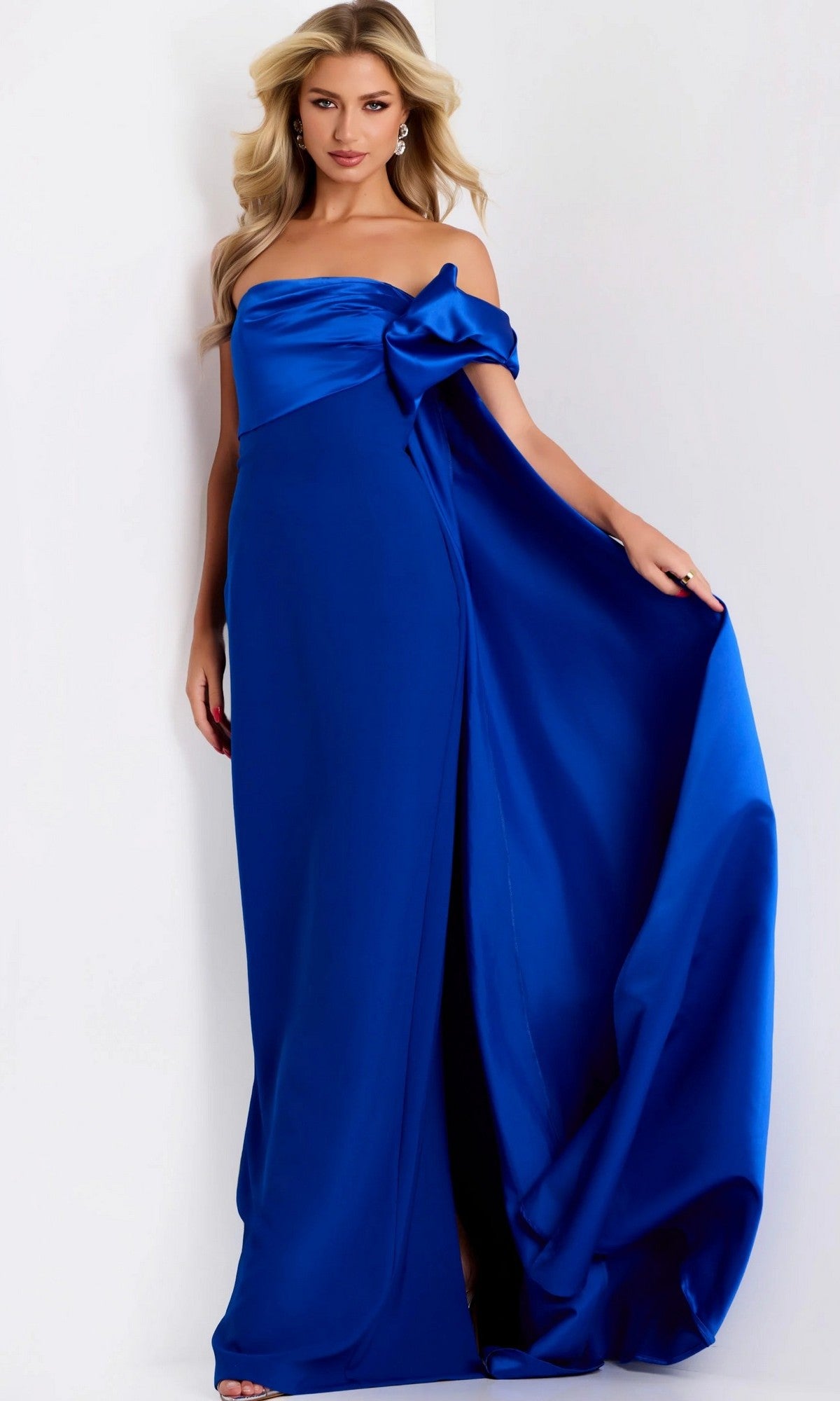A woman wearing the Long Formal Dress: Jovani 38864 in royal blue, featuring a strapless design and a large bow detail, holds out the floor-length skirt against a plain white background.
