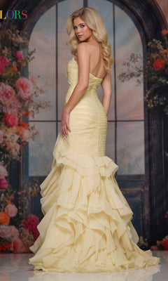 A woman wearing the Strapless Chiffon Prom Gown: Colors Dress 3886 poses before a floral, arched window backdrop, looking over her shoulder in a fitted yellow mermaid gown with a ruffled skirt.