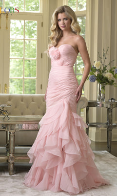 A woman stands indoors near a sofa and large windows, wearing the blush Strapless Chiffon Prom Gown: Colors Dress 3886; flowers decorate a nearby table.