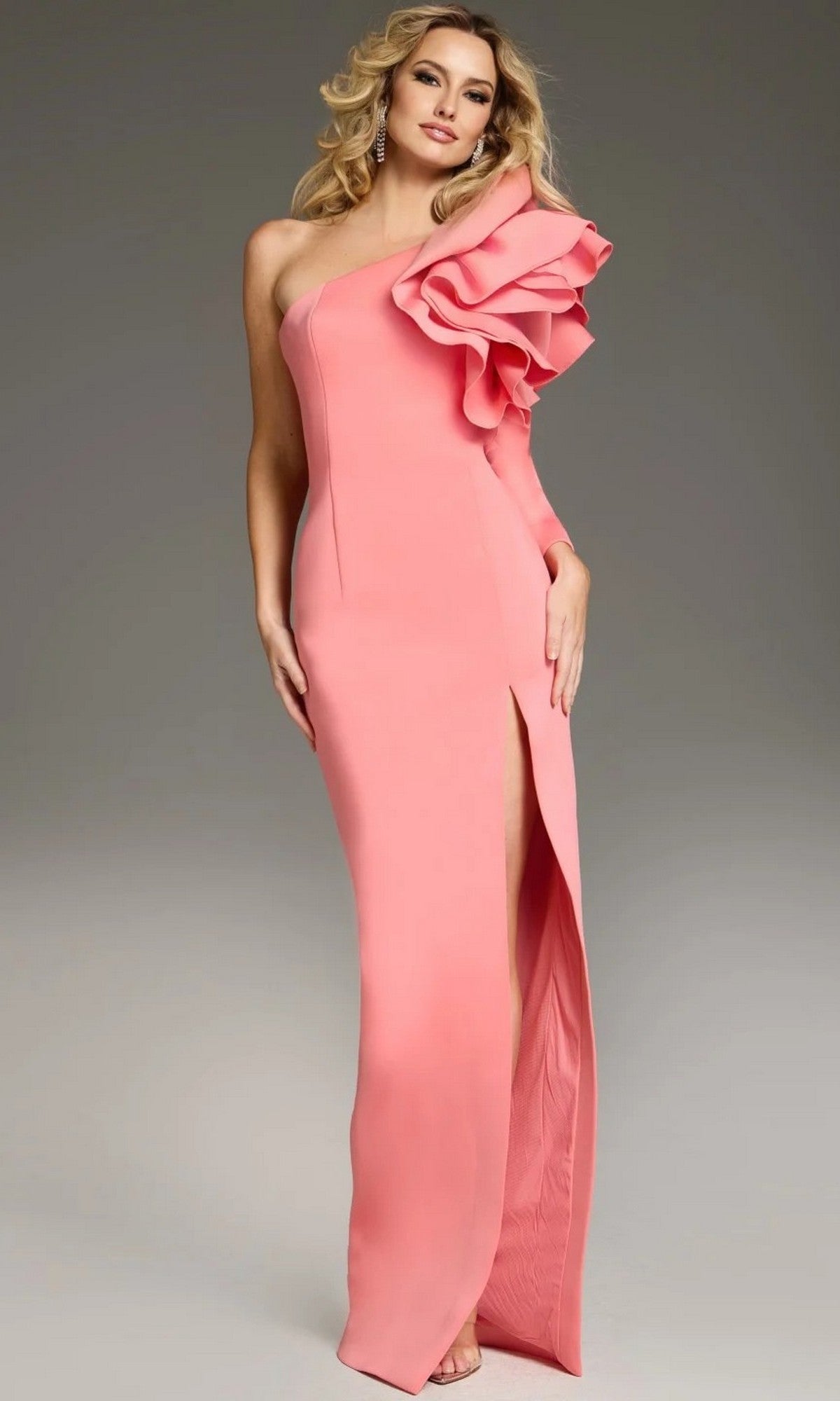 A woman wearing the Jovani 38928 long formal dress—a fitted, floor-length pink designer gown featuring a high-leg slit and a dramatic ruffle detail on one shoulder—stands against a plain background.