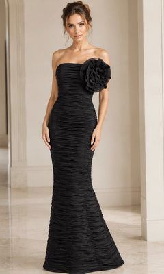 A woman stands indoors wearing the elegant Jovani 38947—a long formal black dress featuring a strapless, floor-length silhouette with ruched fabric and a statement flower accent on the bodice.