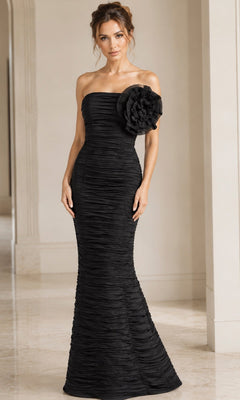 A woman wearing the Jovani 38947 long formal dress—a strapless, floor-length black designer gown featuring ruched fabric and a large floral detail on one side—stands indoors on a marble floor.