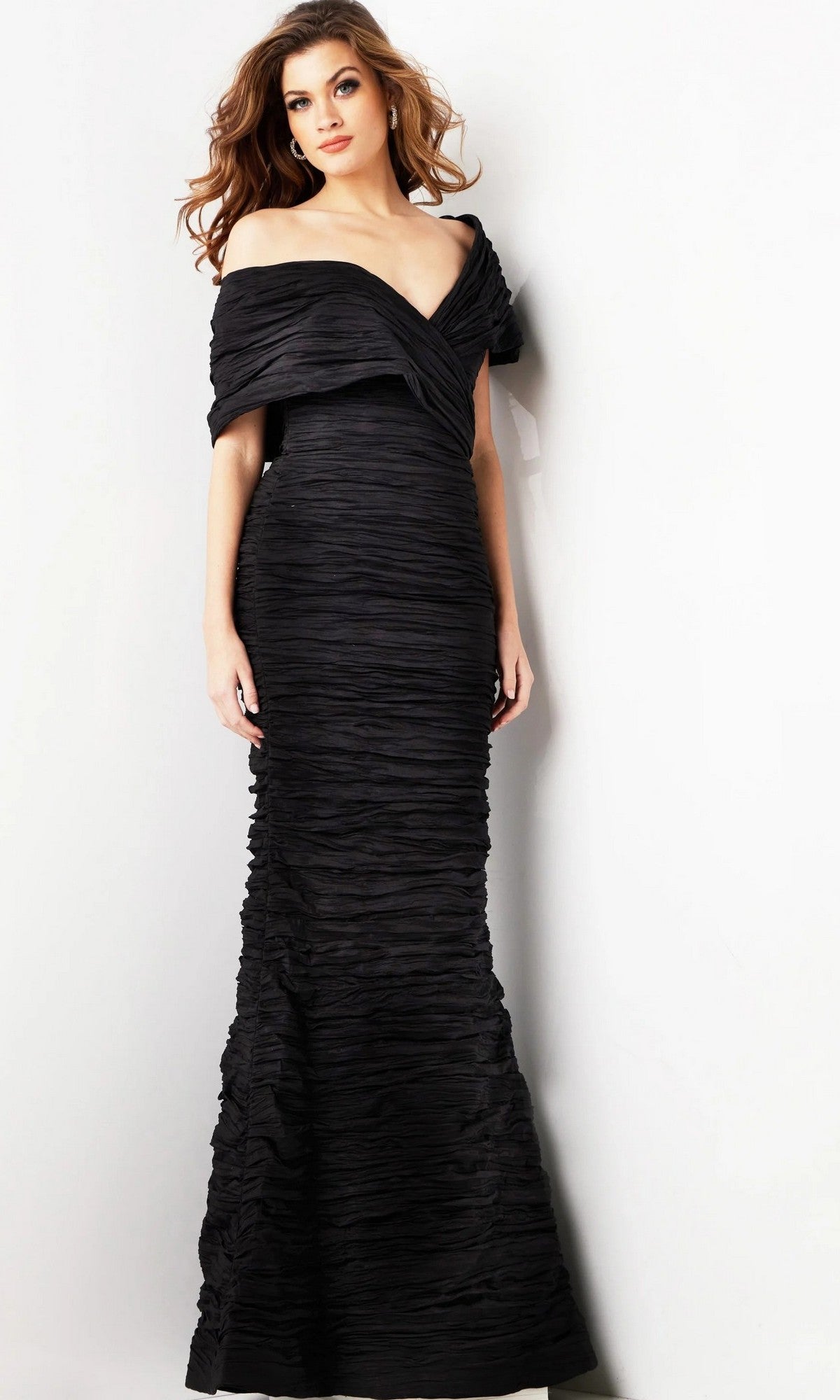 A woman wearing the Jovani 38954 long formal dress in black, which is a floor-length, off-the-shoulder evening gown with ruched fabric, stands against a plain light background. This designer dress features a sophisticated silhouette that highlights the elegance of its fitted sheath style.