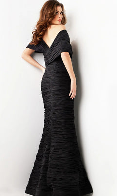 A woman stands sideways against a plain light-colored background, wearing the Jovani 38954 long formal dress—a textured, off-shoulder evening gown with a sleek fitted silhouette reminiscent of high-end designer fashion—while glancing over her shoulder.