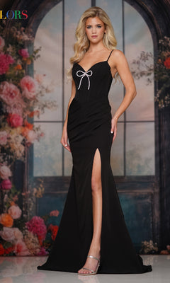 A woman stands indoors before a floral and windowed backdrop, wearing the Colors Dress 3901—a black spaghetti-strap long prom dress with a thigh-high slit and bow detail.