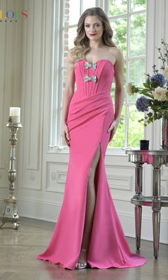 A woman stands indoors by a window and floral table, wearing the Colors Dress 3902—an elegant hot pink strapless long corset prom gown with a thigh-high slit and bow details.