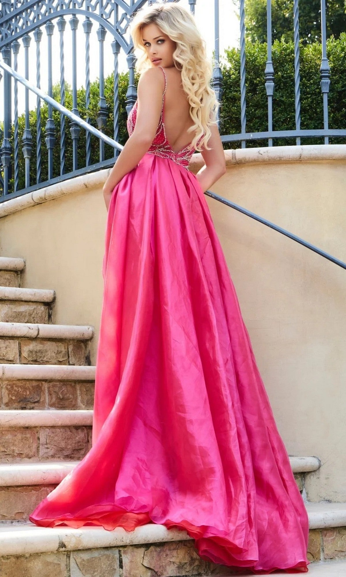 A woman with long blonde hair wears the Long Formal Dress: Jovani 39042 in bright pink as she stands on stone steps, glancing over her shoulder, with an iron fence and lush greenery behind her.