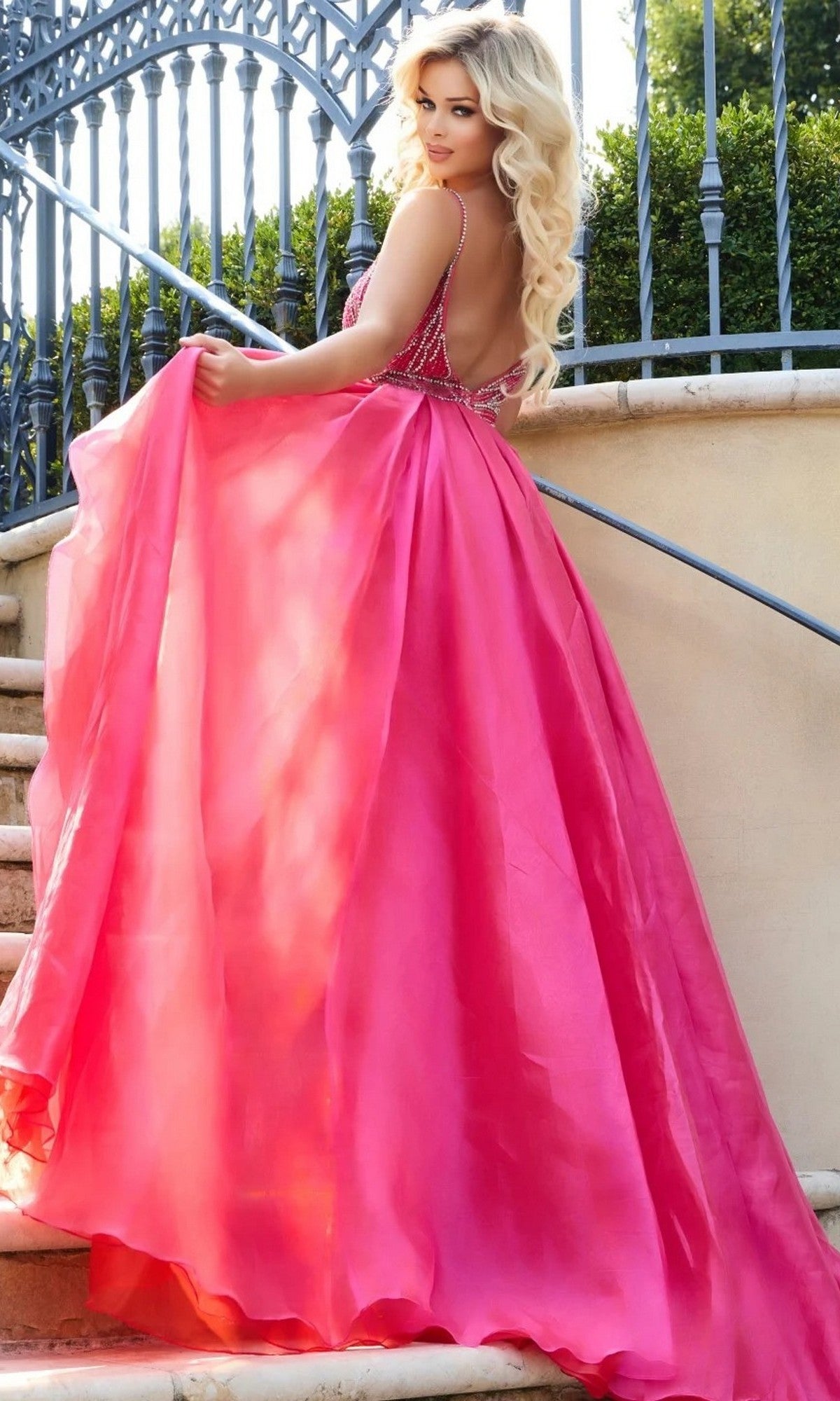 A woman with long blonde hair wears the Long Formal Dress: Jovani 39042, a backless bright pink gown, as she stands on outdoor stairs looking over her shoulder, with a wrought iron fence and greenery in the background.