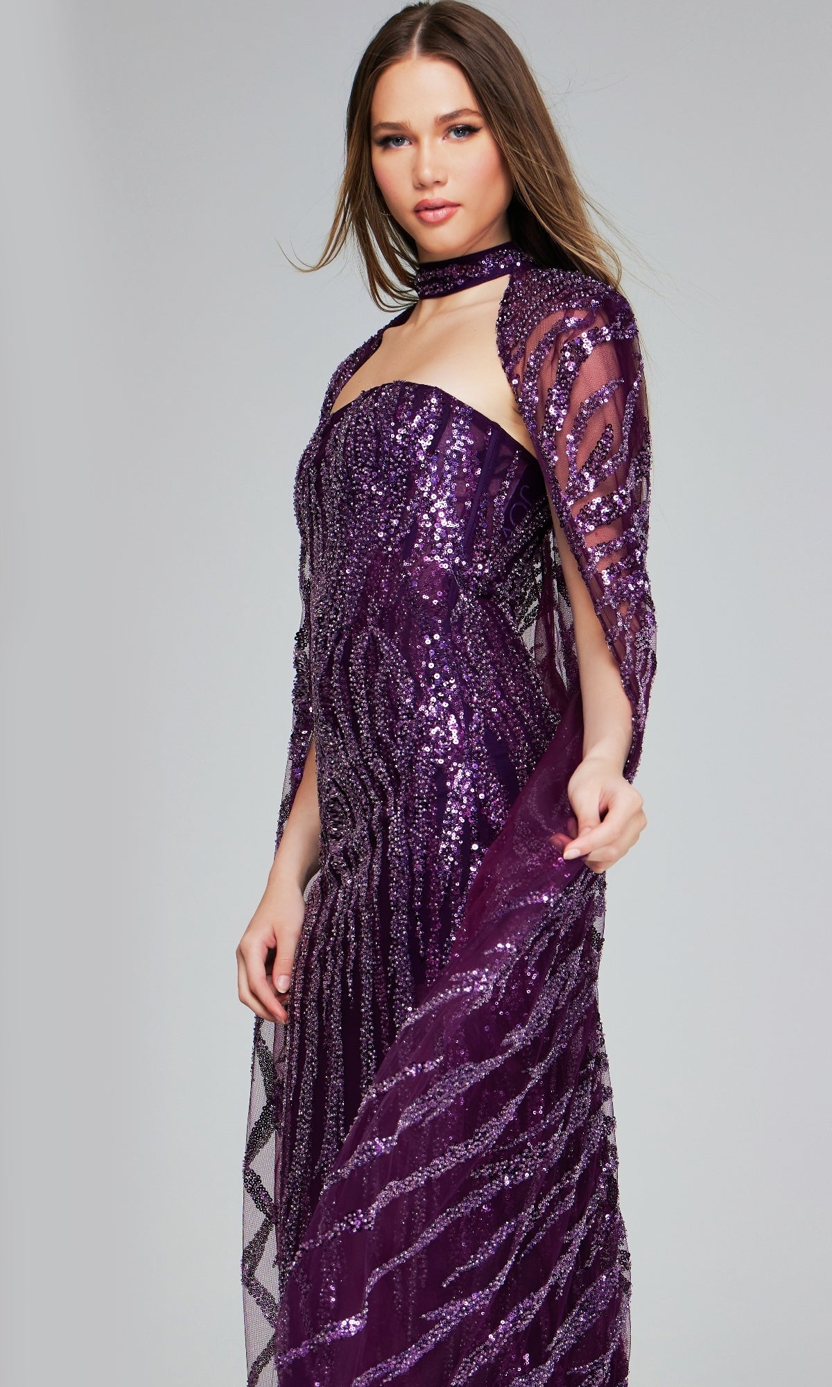 A woman models the Jovani 39046 Purple Sequin Pageant Dress with a matching sheer cape and choker, standing against a plain background—an ideal choice for any special occasion.
