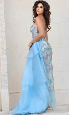 A woman with long dark hair stands against a light stone wall, looking over her shoulder while wearing the Jovani 39055 long formal dress—a shimmering blue and silver beaded mesh gown featuring a layered train.