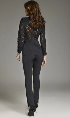A woman stands facing away, wearing the Long Jumpsuit: Jovani 39074, featuring lace sleeves and a lace back, paired with black heels. Her hair is styled in loose waves against a plain, neutral background.