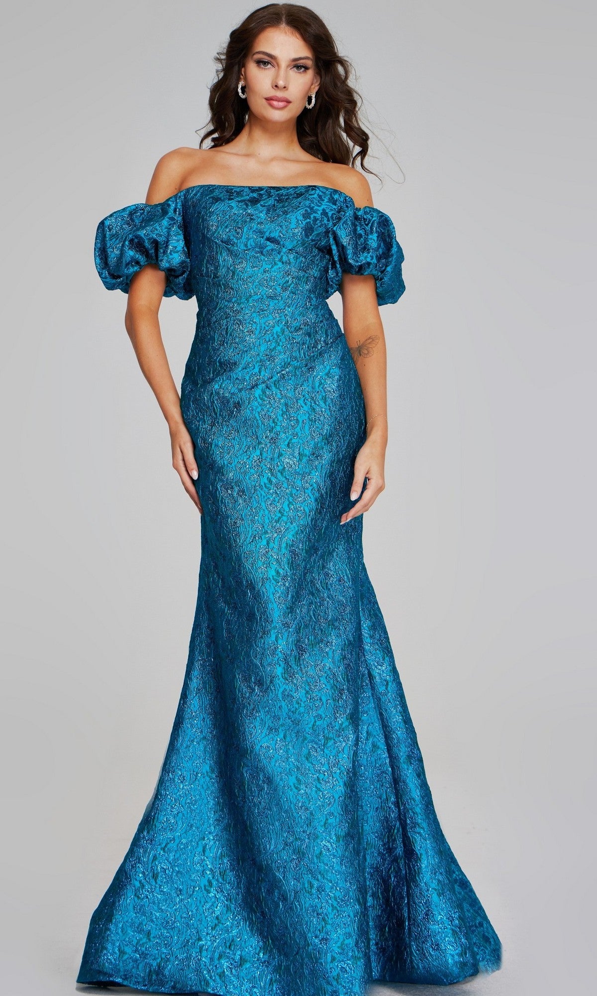 A woman stands against a plain light gray background wearing the Jovani 39113 long formal dress—a teal, off-the-shoulder, floor-length metallic jacquard gown featuring puffed sleeves and a subtle mermaid silhouette.