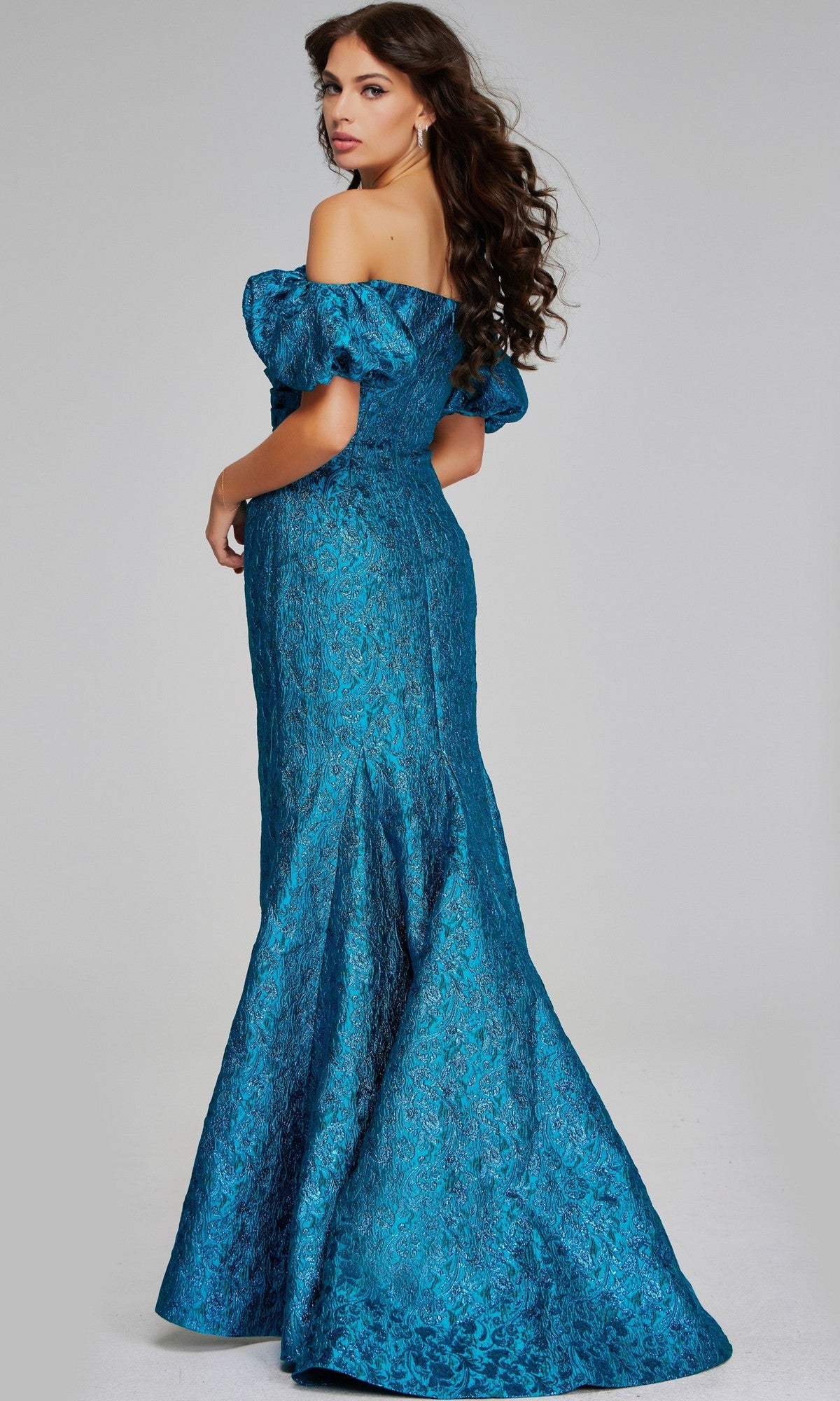 A woman wearing the Jovani 39113 long formal dress, featuring an off-the-shoulder neckline, floor-length metallic jacquard fabric, a fitted bodice, and a subtle mermaid silhouette, stands against a plain light background—an exquisite choice for any elegant occasion.