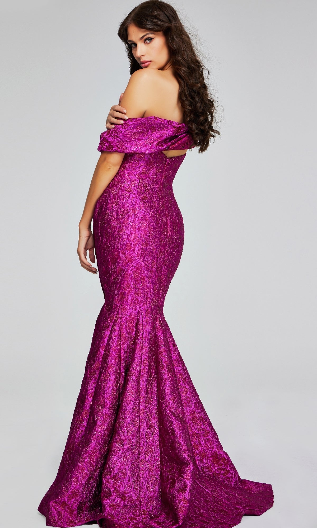 A woman wears the Jovani 39114 long formal dress, a fitted off-the-shoulder magenta mermaid gown with a textured pattern. She stands facing backward, elegantly glancing over her shoulder.