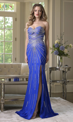 A woman wears the Colors Dress 3921, a strapless royal blue prom dress with hot-stone embellishments, fitted silhouette, and thigh-high slit, standing indoors near a window and floral display.