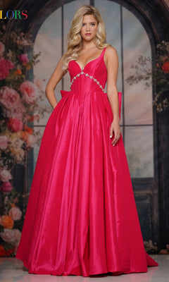 A woman with long blonde hair wears the Colors Dress 3922 Statement-Bow Long Prom Dress by Colors Dress, a bright pink A-line gown with a fitted bodice and jeweled waist, standing before a floral and arched window backdrop.