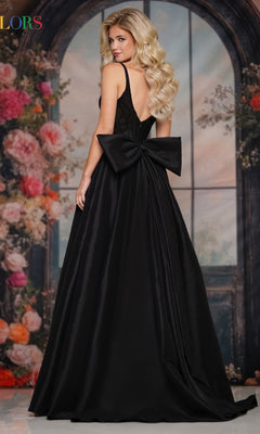 A woman with long, wavy blonde hair models the Colors Dress Statement-Bow Long Prom Dress (Style 3922), a sleeveless black gown with a deep V-back and large waist bow, posing before floral décor and an arched window.