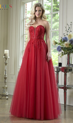 A woman stands indoors wearing the Strapless Long Corset Prom Dress: Colors Dress 3923, featuring a fitted bodice and tulle skirt in red; flowers and a window appear in the background.