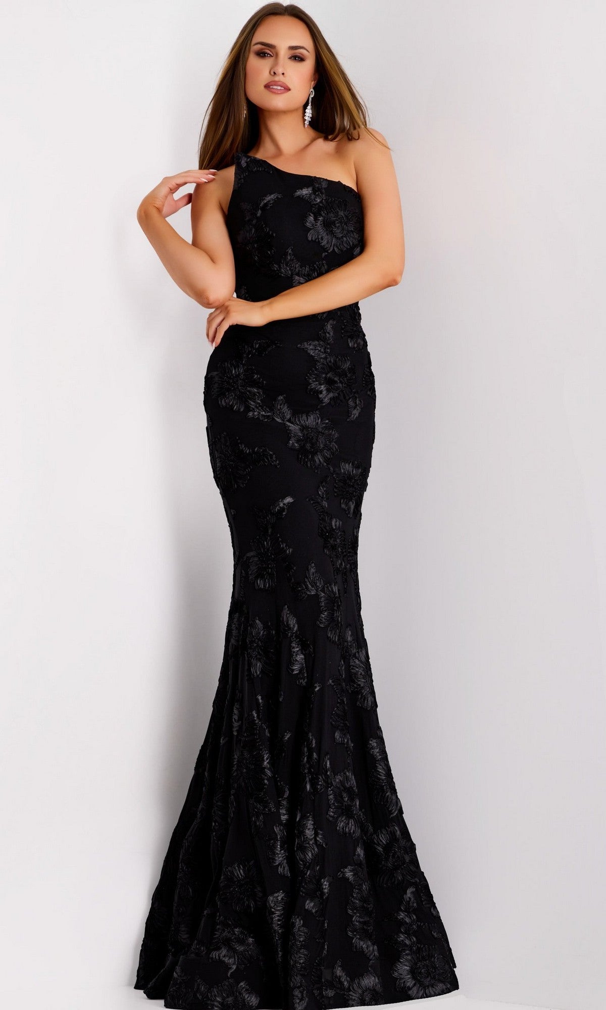 A woman poses against a plain white background wearing the Long Formal Dress: Jovani 39267 in black, a floor-length, one-shoulder designer gown featuring a subtle floral pattern.