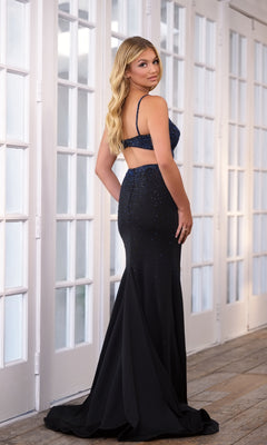 A woman stands indoors wearing the Long Prom Dress 39284 by Ava Presley—a fitted, floor-length black gown featuring a beaded top, open back, and v-neckline—while looking over her shoulder.