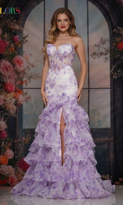 A woman stands before a floral, arched window backdrop wearing the Colors Dress 3928—a strapless lavender prom gown with a floral print, fitted bodice, ruffled skirt, and front slit.