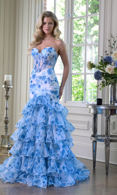 A woman wearing the Strapless Long Print Prom Dress: Colors Dress 3928 stands by large windows in a beautifully decorated room.