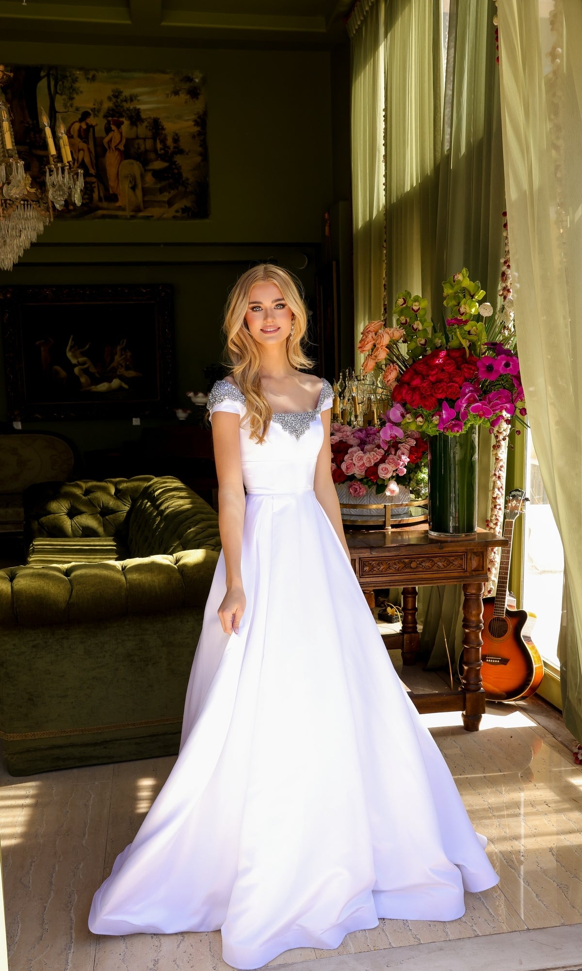 A woman in the Long Prom Dress 39322 by Ava Presley stands in a room with green walls, floral arrangements, a green sofa, paintings, and a guitar by the window.