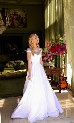 A woman in the Long Prom Dress 39322 by Ava Presley stands in a room with green walls, floral arrangements, a green sofa, paintings, and a guitar by the window.