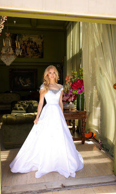 A woman wearing the Ava Presley Long Prom Dress 39322, a white floor-length gown with cap sleeves, stands in a doorway with a green living room and floral arrangements visible in the background.