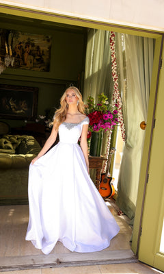 A woman in the Long Prom Dress 39322 by Ava Presley stands in a doorway, holding her elegant a-line gown. Behind her are a green sofa, flowers, a guitar, and framed artwork.