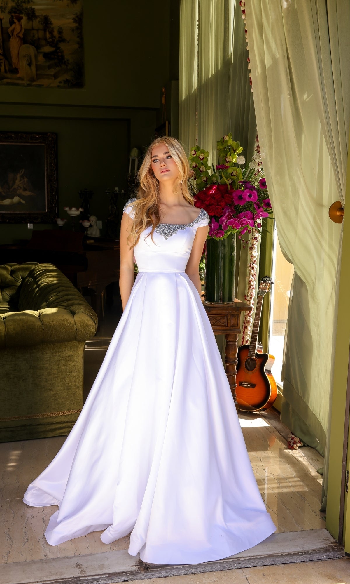 A woman in the Long Prom Dress 39322 by Ava Presley stands indoors near a large window, with a bouquet of flowers, a guitar, and a green sofa in the background.