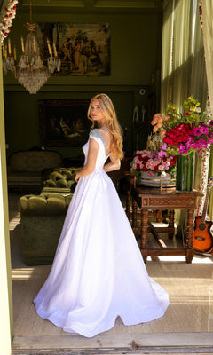 A woman wearing the Long Prom Dress 39322 by Ava Presley, a white A-line ball gown with silver details and cap sleeves, stands indoors among vintage decor, flowers, and a guitar near a window with green curtains.