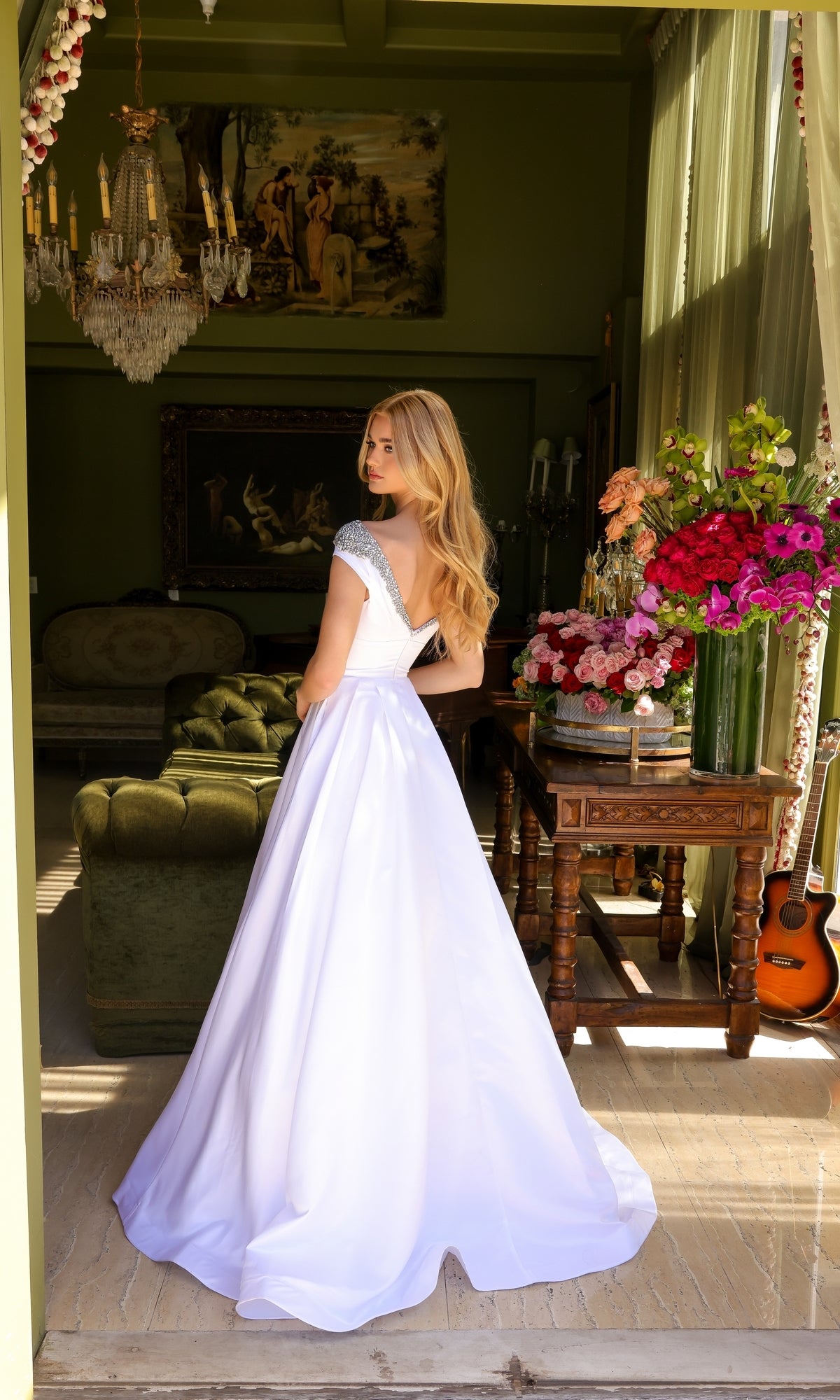 A woman in the Long Prom Dress 39322 by Ava Presley, featuring an a-line silhouette and beaded neckline, stands indoors among green furniture, floral arrangements, a guitar, and framed artwork.