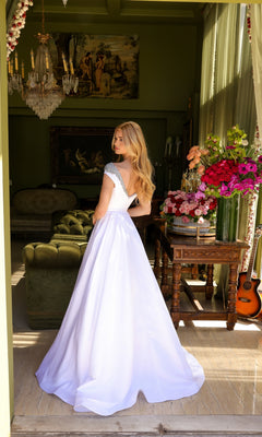 A woman in the Long Prom Dress 39322 by Ava Presley, featuring an a-line silhouette and beaded neckline, stands indoors among green furniture, floral arrangements, a guitar, and framed artwork.