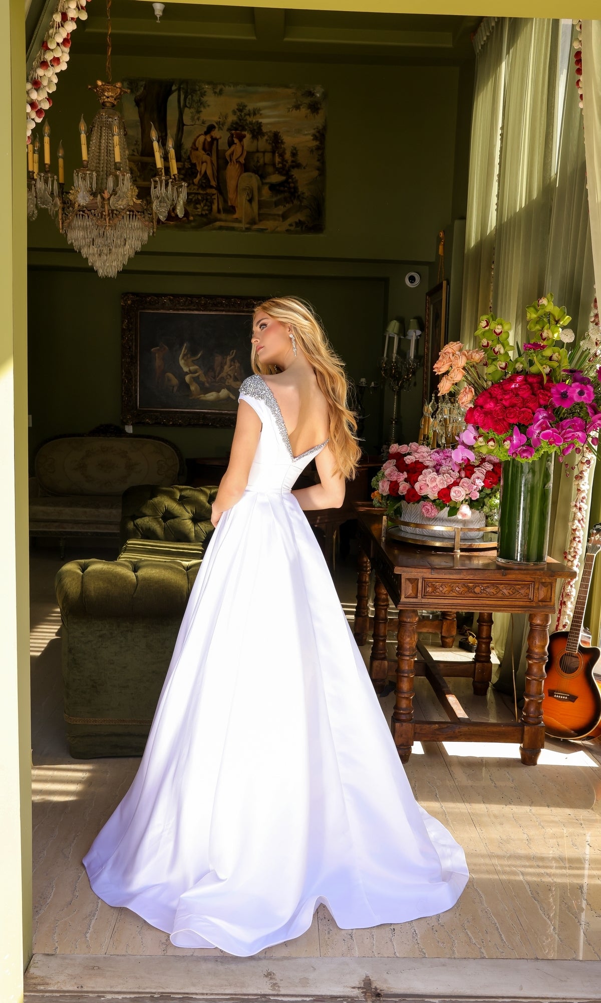 A woman wearing the Long Prom Dress 39322 by Ava Presley, a white backless formal gown with cap sleeves, stands indoors near a window, with flowers, a sofa, and a guitar in the background.