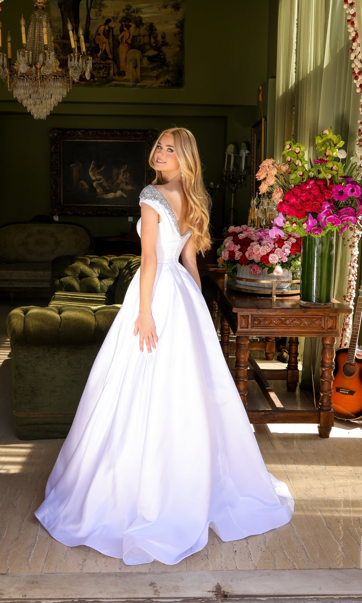 A woman in the Long Prom Dress 39322 by Ava Presley, an elegant a-line ball gown with cap sleeves, stands in a room filled with vintage furniture, holding a bouquet of flowers near a window with a guitar beside her.