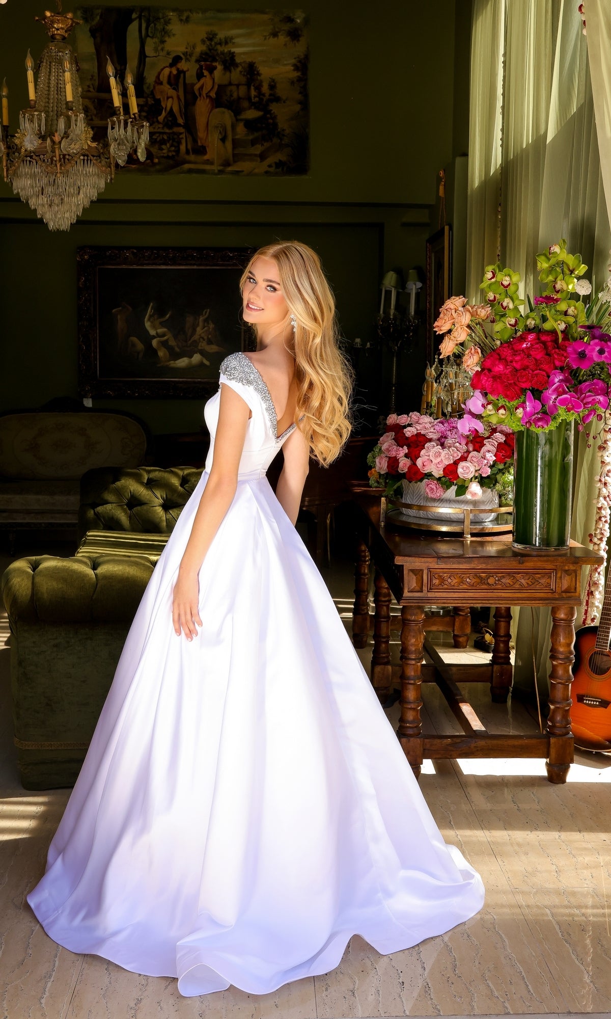 A woman wears the Long Prom Dress 39322 by Ava Presley, a white A-line ball gown with cap sleeves, standing indoors near a table of colorful flowers in a room with classic décor and paintings.