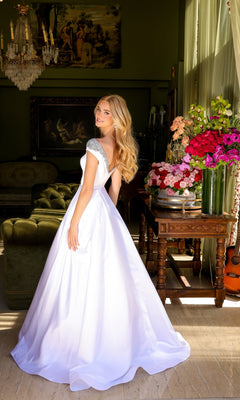 A woman wears the Long Prom Dress 39322 by Ava Presley, a white A-line ball gown with cap sleeves, standing indoors near a table of colorful flowers in a room with classic décor and paintings.