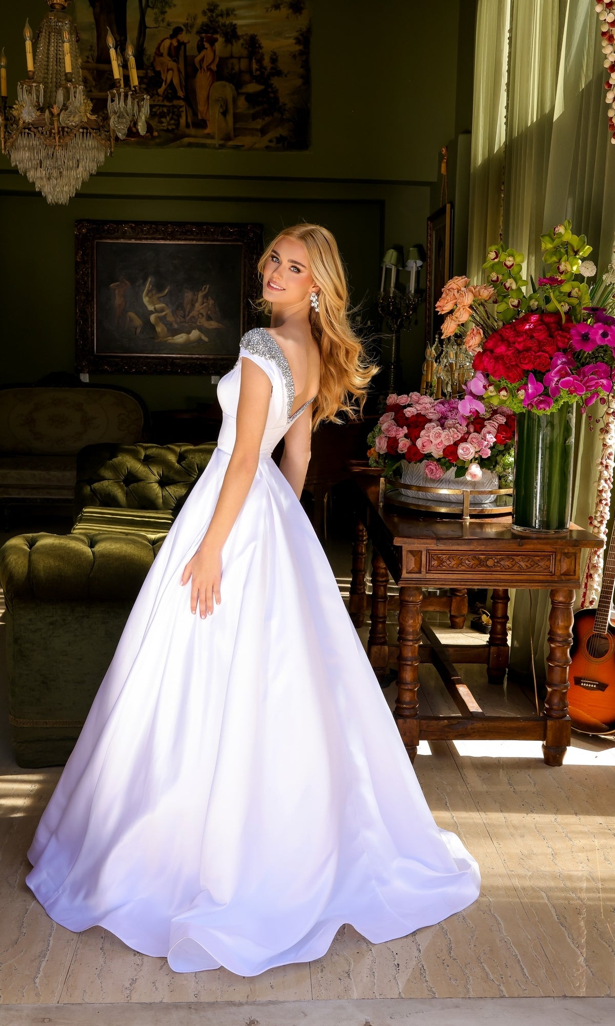 A woman wearing the Long Prom Dress 39322 by Ava Presley, a white A-line ball gown with embellished shoulders, stands indoors near a table with colorful flowers, looking back over her shoulder.