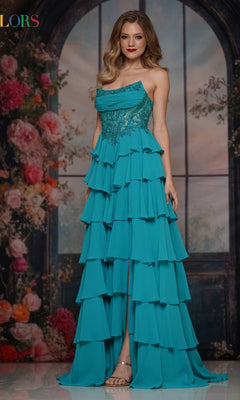A woman in the Colors Dress 3934, a strapless long ruffled prom dress, stands indoors before a floral backdrop with loose waves and open-toed heels completing her formal look.