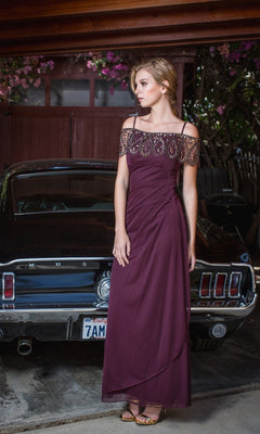 A woman in a burgundy Off-Shoulder Draped Long Formal Dress: Eureka 3937 poses by a classic black Ford Mustang in a garage adorned with flowers and greenery, dressed for a black-tie event.
