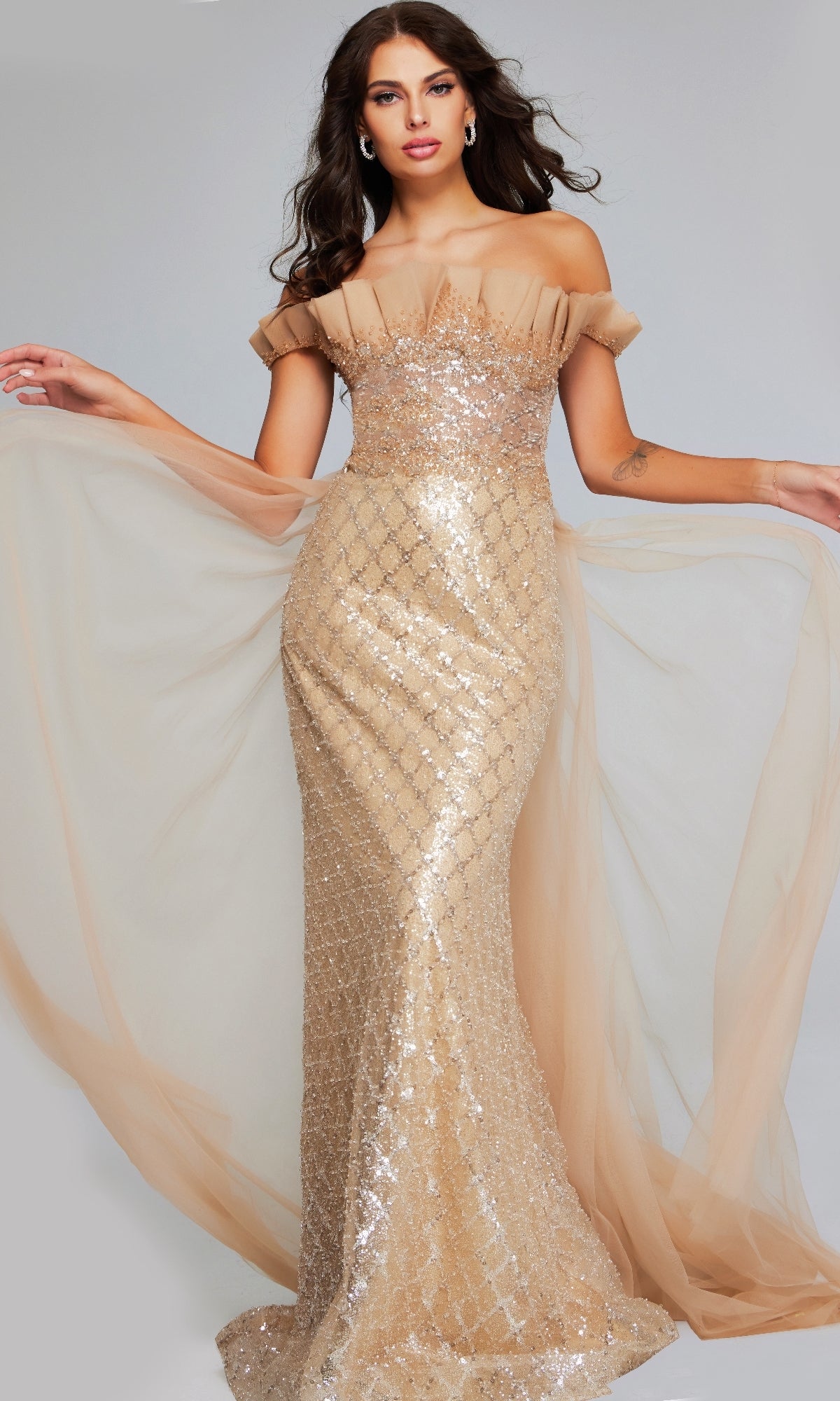A woman stands with arms slightly raised, wearing the Jovani 39389 long formal dress—an off-the-shoulder sequin evening gown with sheer tulle accents.