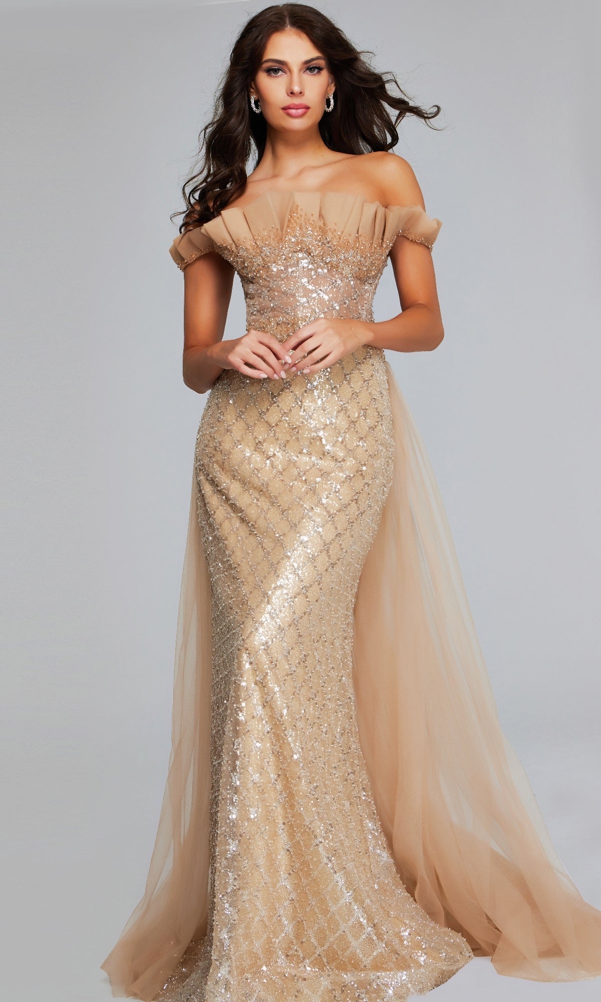 A woman stands against a plain light gray background wearing the Jovani 39389 Long Formal Dress, a floor-length, off-the-shoulder mermaid gown with gold sequins and tulle accents.