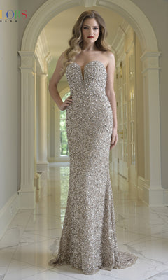 A woman stands in a hallway wearing the Colors Dress 3939, a floor-length, strapless prom dress with silver sequins and a deep neckline.